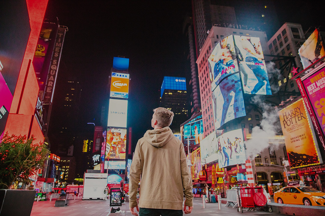 Man in a tan hoodie facing advertisements in Times Square